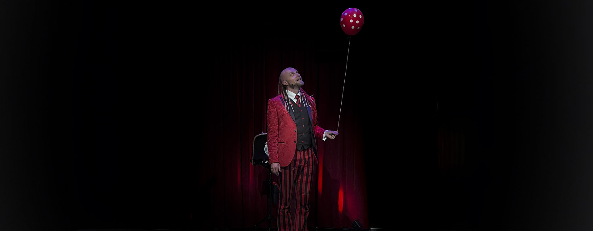 Trollkarlen Arkadia står klädd i rött med en röd ballong i handen.